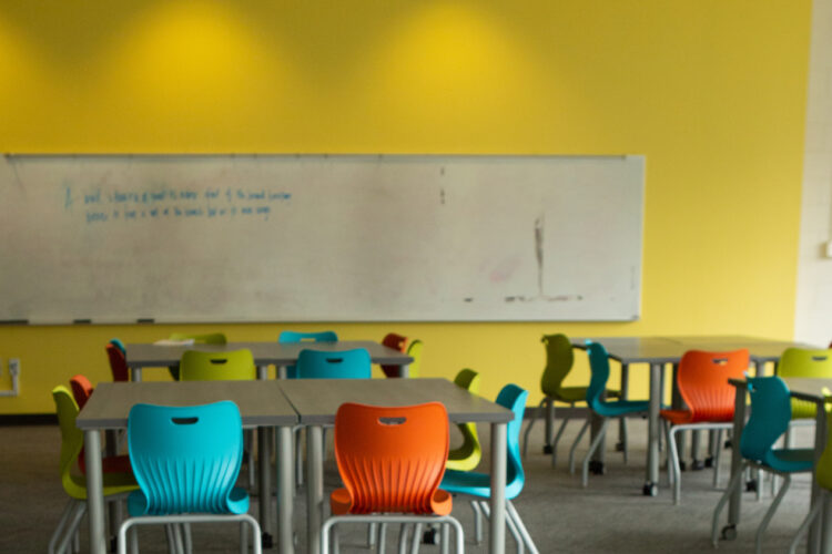 An empty classroom with unoccupied tables and chairs with a white dry erase board attached to a yellow wall.