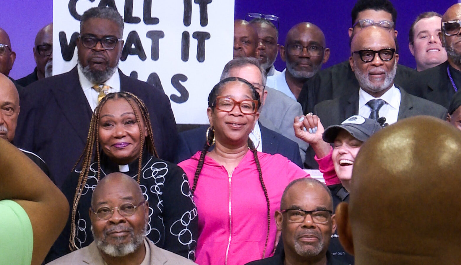 Attendees of the gathering pose for a group photo. A Black woman in the foreground holds her fist up. Behind her and several others is a sign that says "Call it what it was".