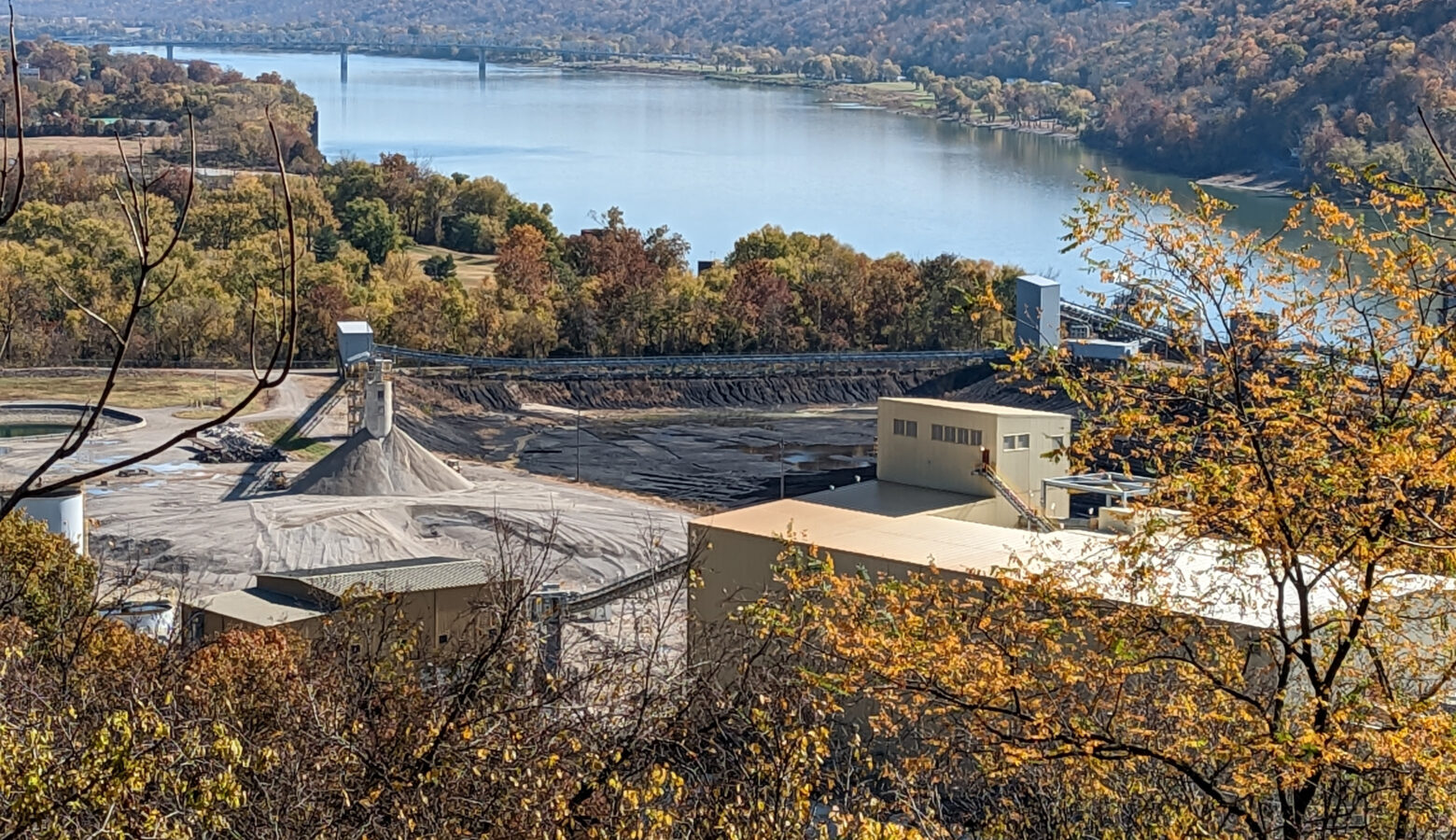 A coal ash pond at the Clifty Creek coal plant in Jefferson County. Bare dirt sits in front of a yellow building. In the background are trees surrounding a body of water with a bridge crossing it.