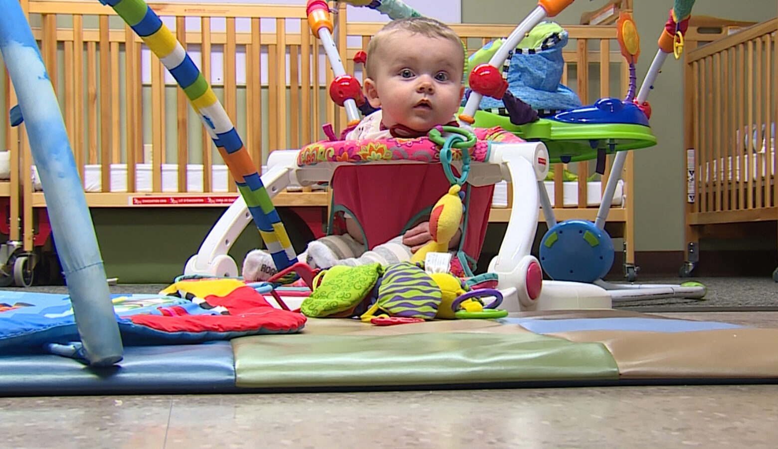 A toddler sits inside a colorful baby bouncer inside a room with cribs in the background.