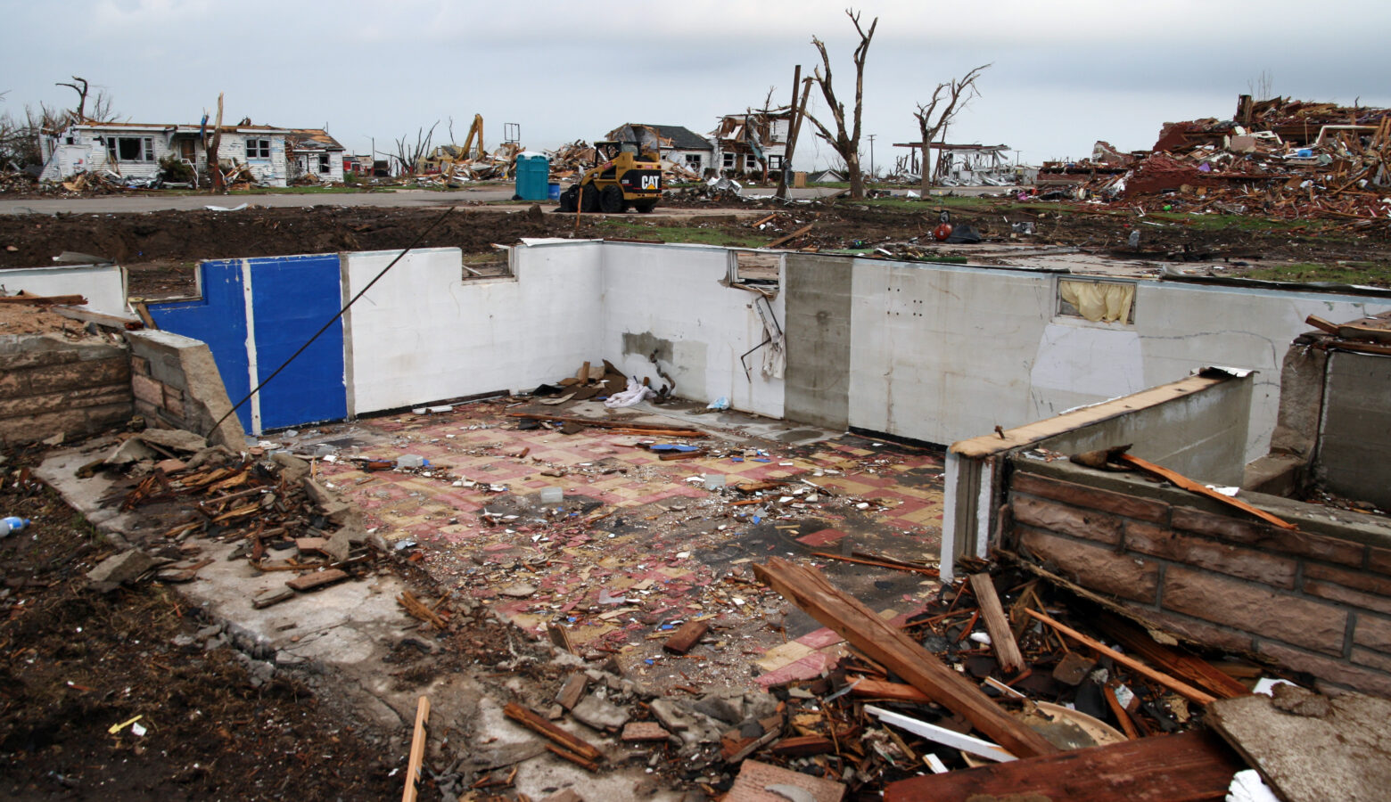 Debris is strewn across a flat landscape from where a home was demolished by a tornado. All that is left of the home is its basement. Across the street, other homes are damaged or partially collapsed. Construction equipment is parked on the street as the cleanup effort has already begun to collect the debris.