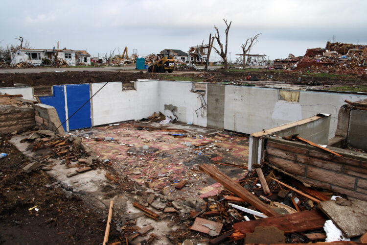 Debris is strewn across a flat landscape from where a home was demolished by a tornado. All that is left of the home is its basement. Across the street, other homes are damaged or partially collapsed. Construction equipment is parked on the street as the cleanup effort has already begun to collect the debris.