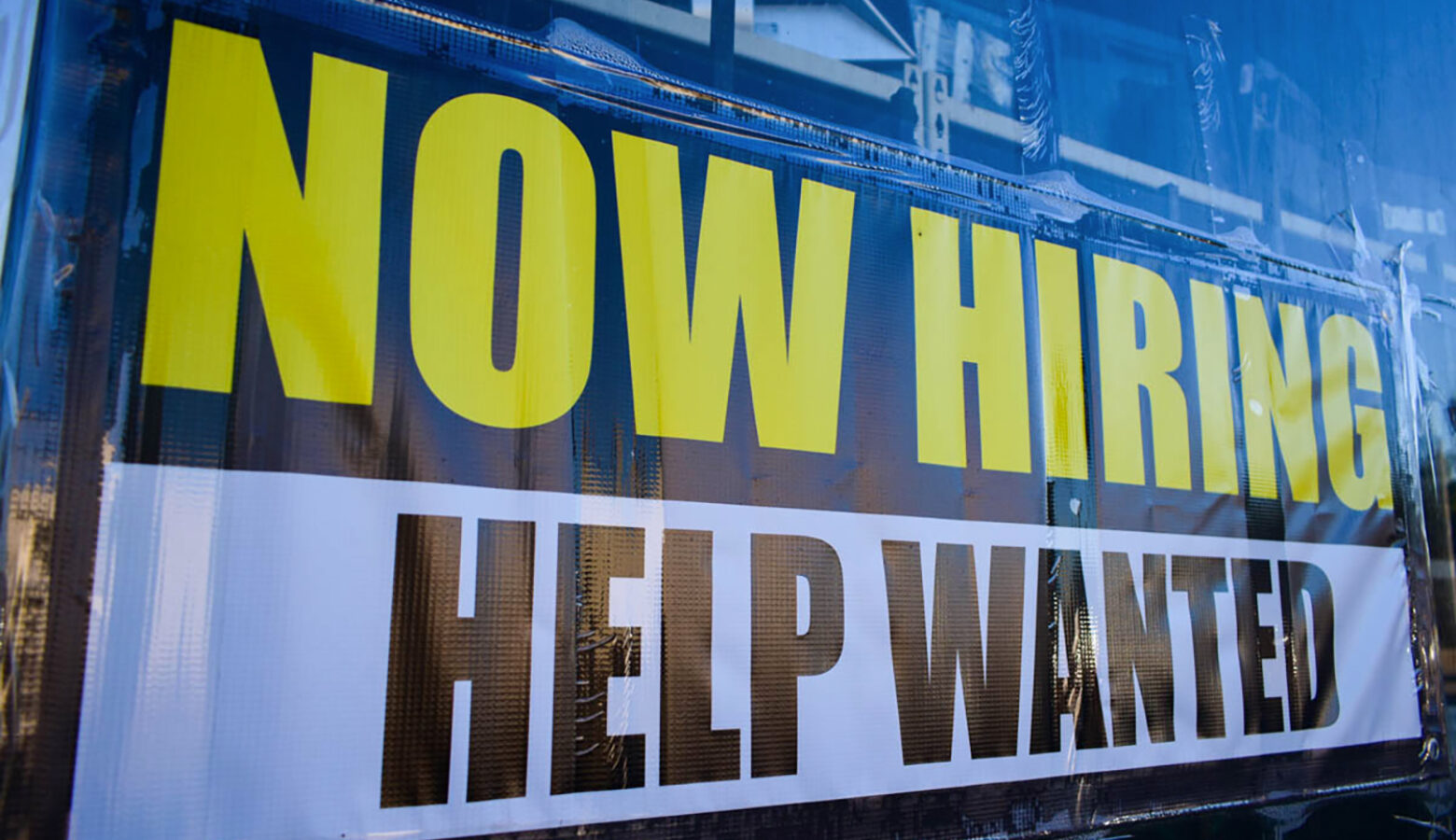 A sign reads "NOW HIRING" in bold yellow letters and "HELP WANTED" in bold black letters underneath.