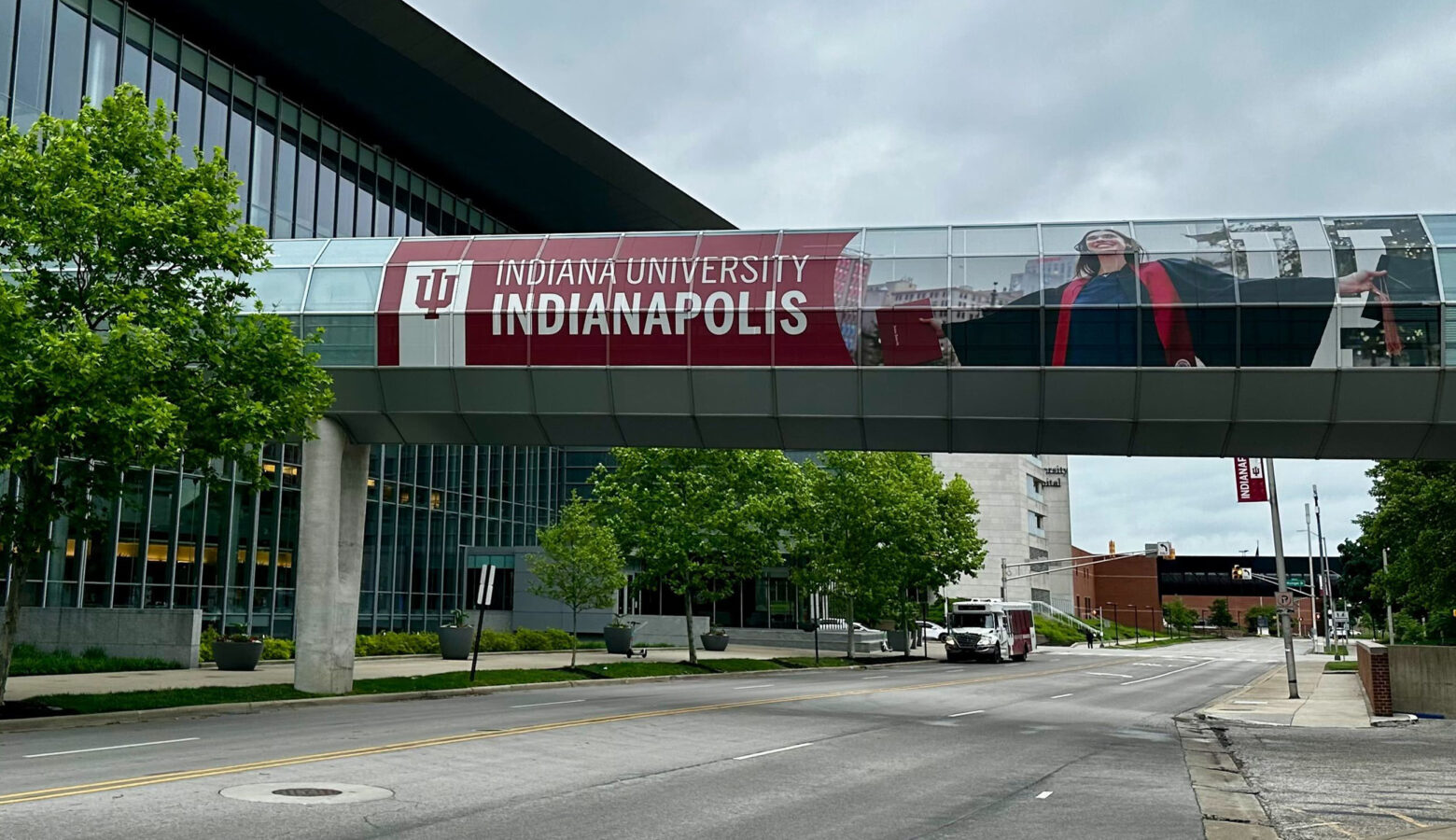 A street scene at the IU Indianapolis campus with an elevated walkway attaching two buildings. The walkway features an "Indiana University Indianapolis" sign and photo of a female graduate in a robe holding a red diploma.