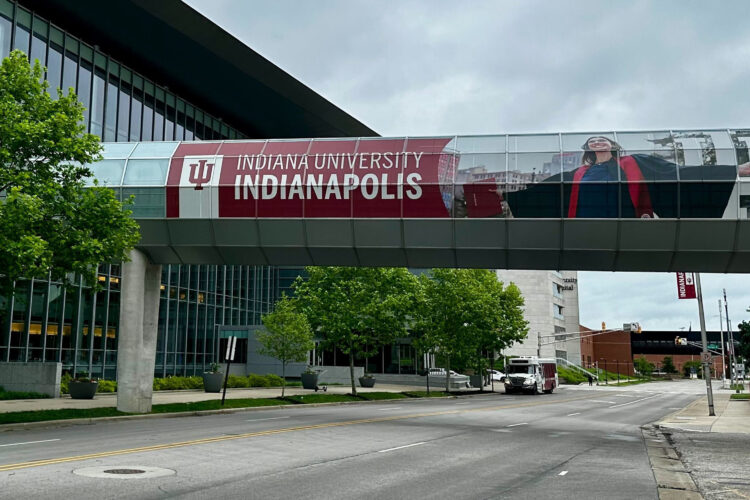 A street scene at the IU Indianapolis campus with an elevated walkway attaching two buildings. The walkway features an "Indiana University Indianapolis" sign and photo of a female graduate in a robe holding a red diploma.