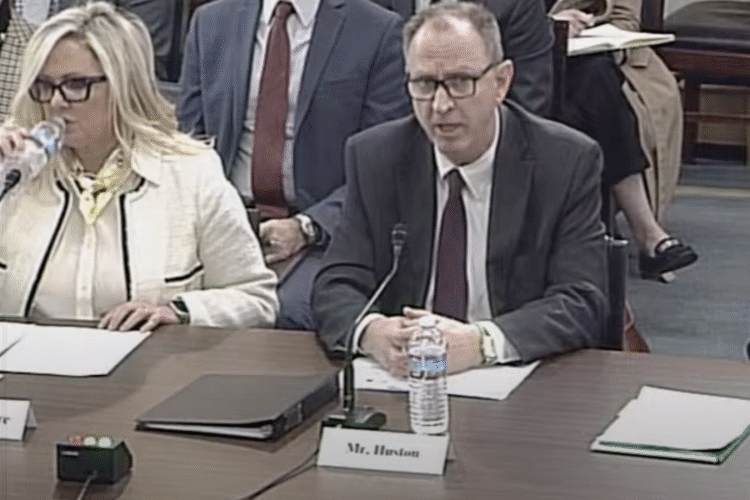 IURC Chairman Jim Huston speaks at a U.S. House Committee on Energy and Commerce hearing on the challenges state utility regulators face in delivering reliable and affordable energy. He is a White man, wearing glasses and a suit. He sits at a table with papers, microphone, water bottle and a small paper name plate that says Mr. Huston.