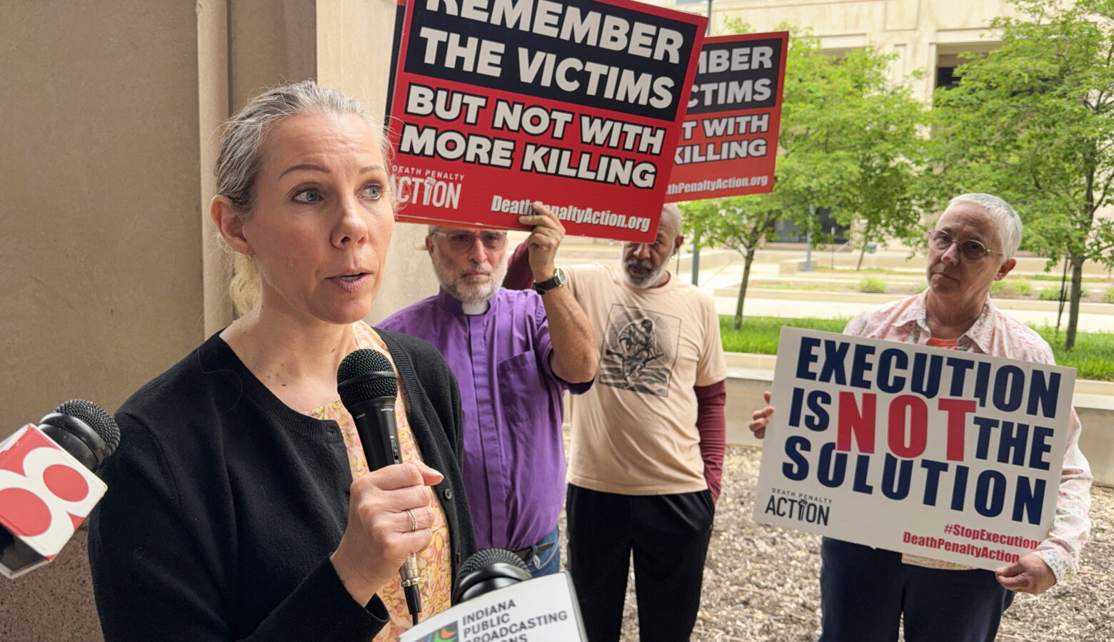 Megan Carter speaks into a microphone while advocates hold up signs behind her that read "Remember the victims but not with more killing" and "Execution is not the solution." Carter is a White woman with blonde hair. She is wearing a black sweater over a yellow top.