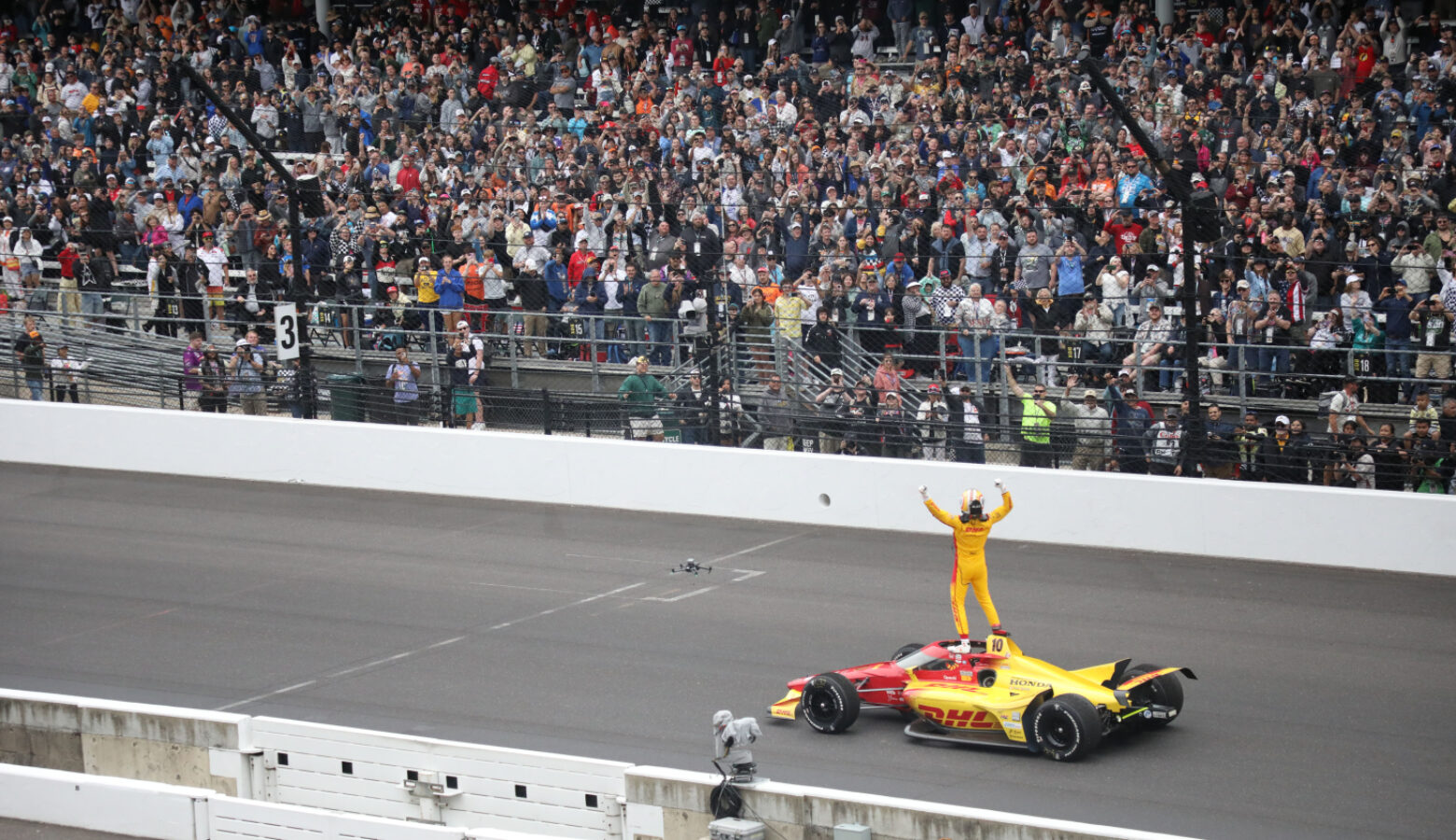 Alex Palou stopped his race car near the yard of bricks finish line at the Indianapolis Motor Speedway. He stands atop his red and yellow car in his yellow and red safety suit pumping both hands in the air to a capacity crowd.