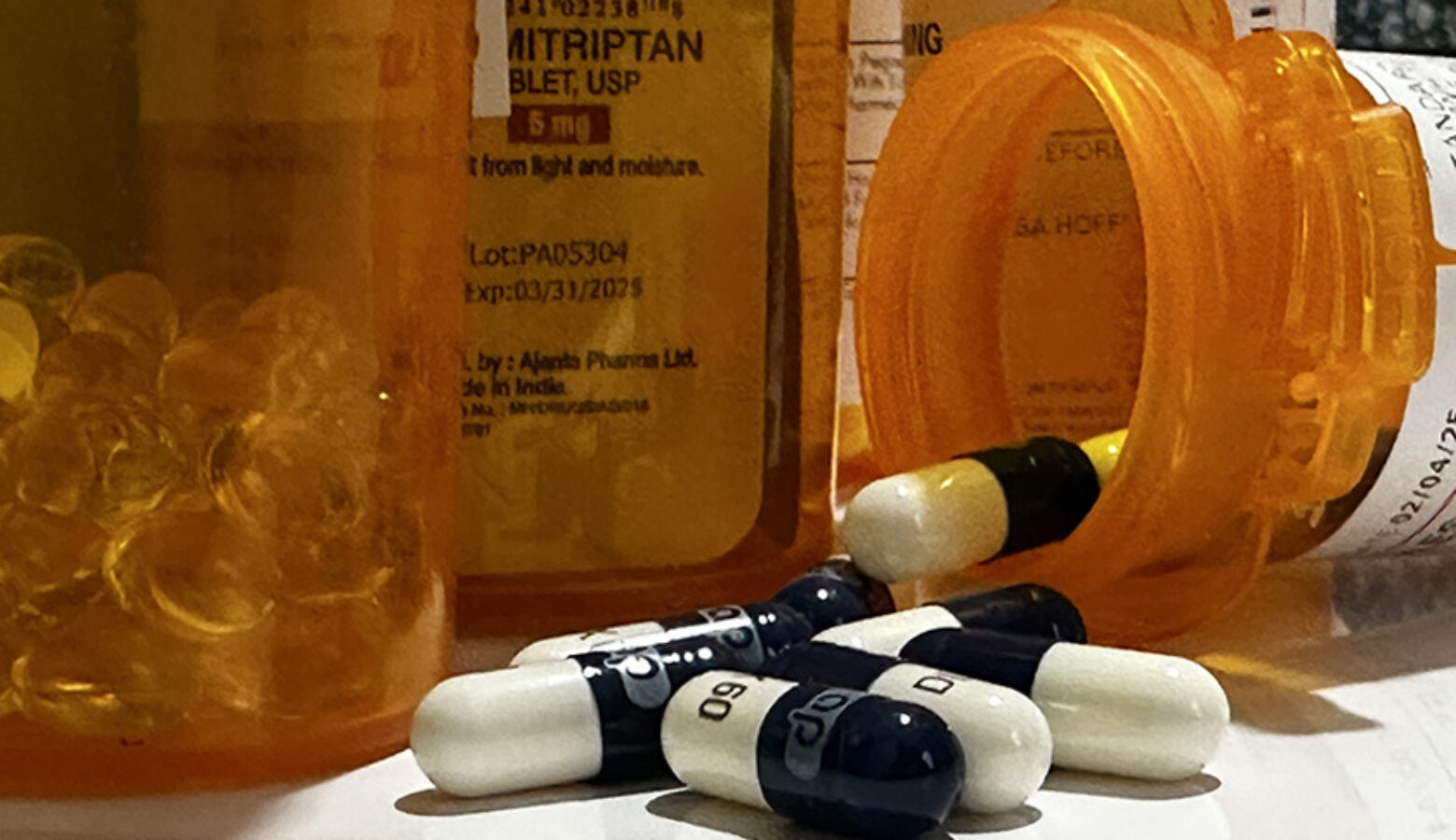 Several prescription pill bottles sit on a white counter top. One rests on its side with white and black capsules spilling out of it.