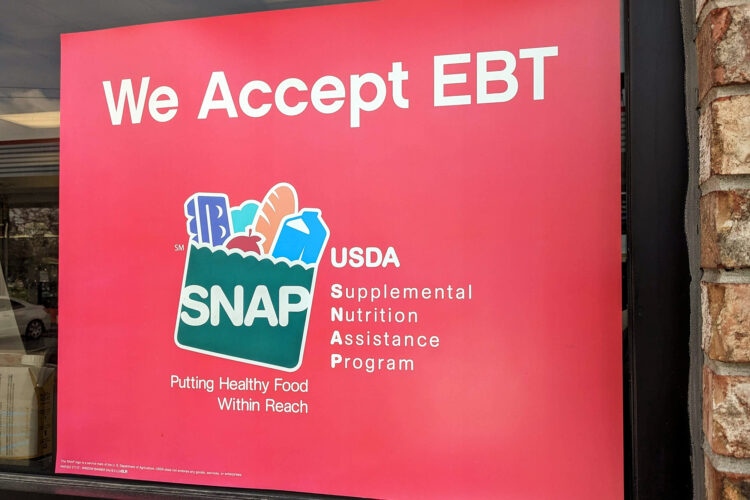 A red window cling is posted on the exterior of brick gas station. It reads "We Accept EBT" across the top. Next to the SNAP logo, which is cartoon paper grocery bag, it says, "USDA, Supplemental Nutrition Assistance Program." Underneath the logo is the program's tagline, "Putting Healthy Food Within Reach."