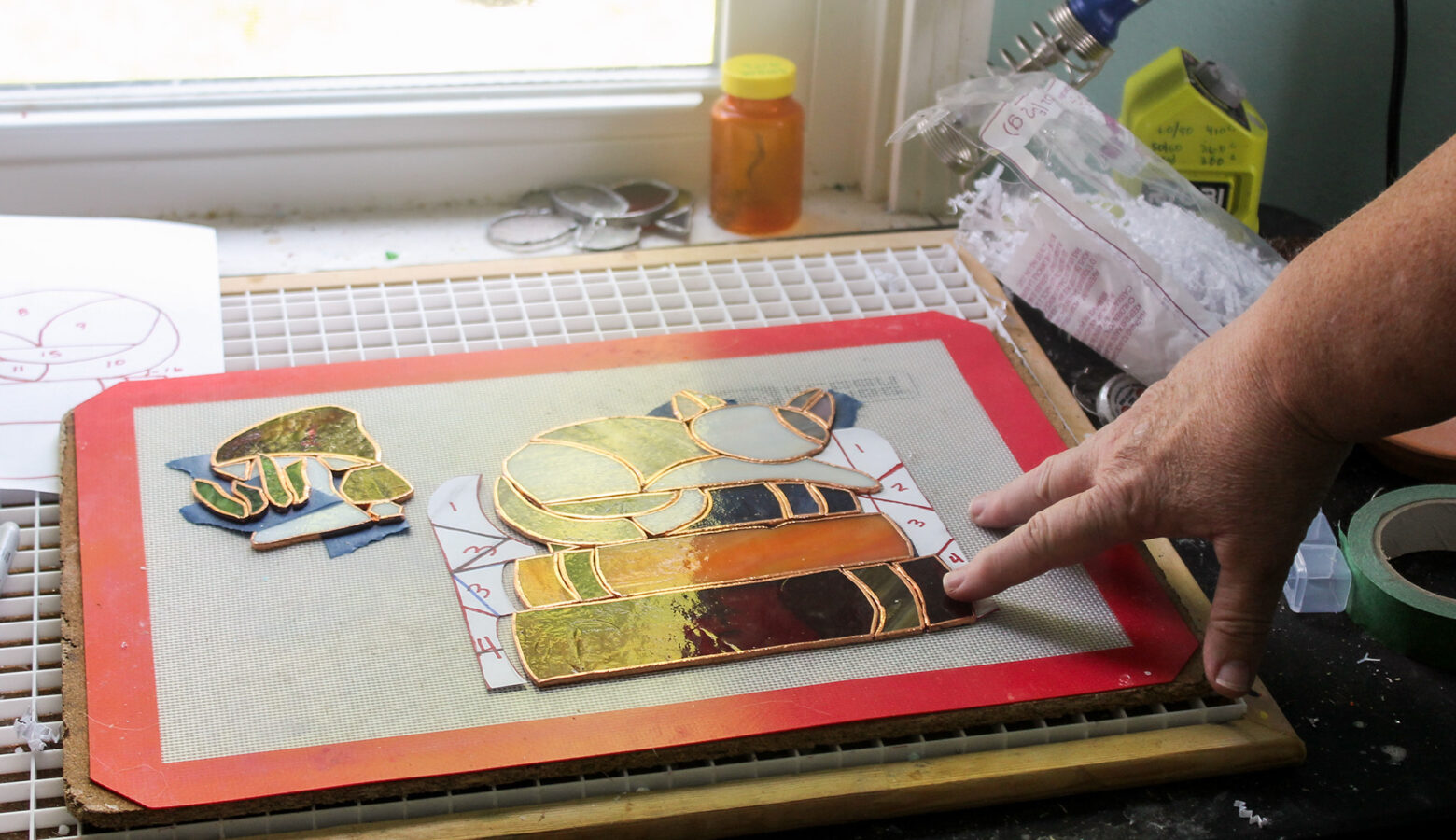 Pat shows her stain glass art of a cat sleeping on books. Her hand touches the edge of a silpat at a workstation next to a window. It's crowded with materials and a pill bottle at the edge of the window pane.