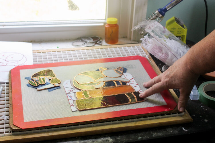 Pat shows her stain glass art of a cat sleeping on books. Her hand touches the edge of a silpat at a workstation next to a window. It's crowded with materials and a pill bottle at the edge of the window pane.
