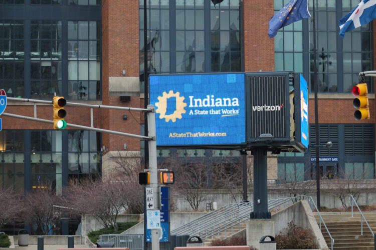 An electronic sign that has a blue background. "Indiana" is written in white letters in the middle of the screen. "A State that works." is written in white underneath "Indiana." A picture of the state is located to the left of the screen outlined in gold.