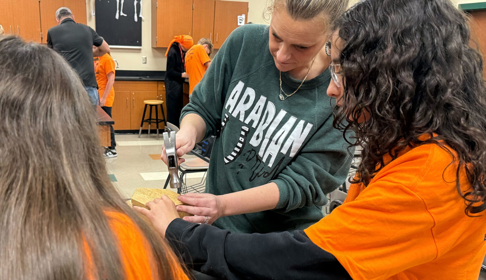 Students wear orange shirts and safety goggles. A woman stands next to the students and wears a green shirt holding a hammer.