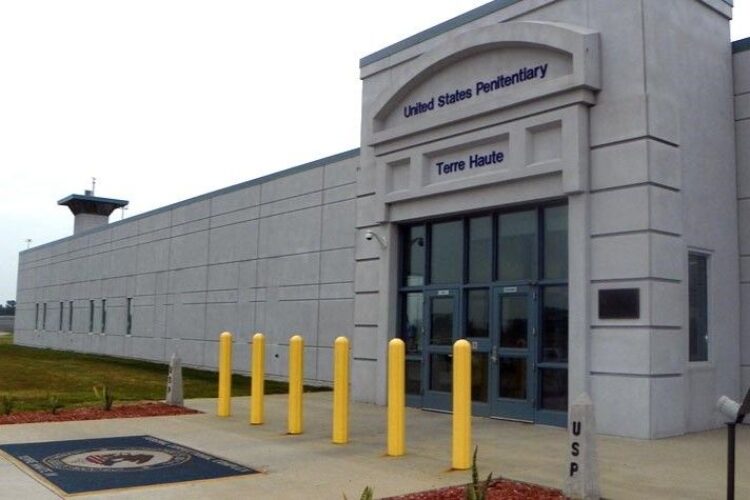 A photo shows the front entrance of the U.S. Prison at Terre Haute. It is a gray concrete structure with six yellow anti-crash posts in front of steel and glass doors.