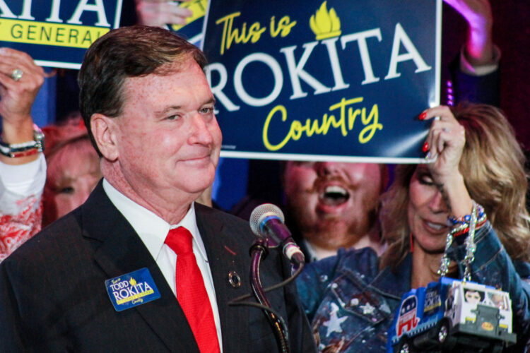 odd Rokita stands in front of supporters who are holding up signs. One sign says "This is Rokita County." Rokita is a White man with dark hair. He is wearing a black suit, white shirt and red tie.