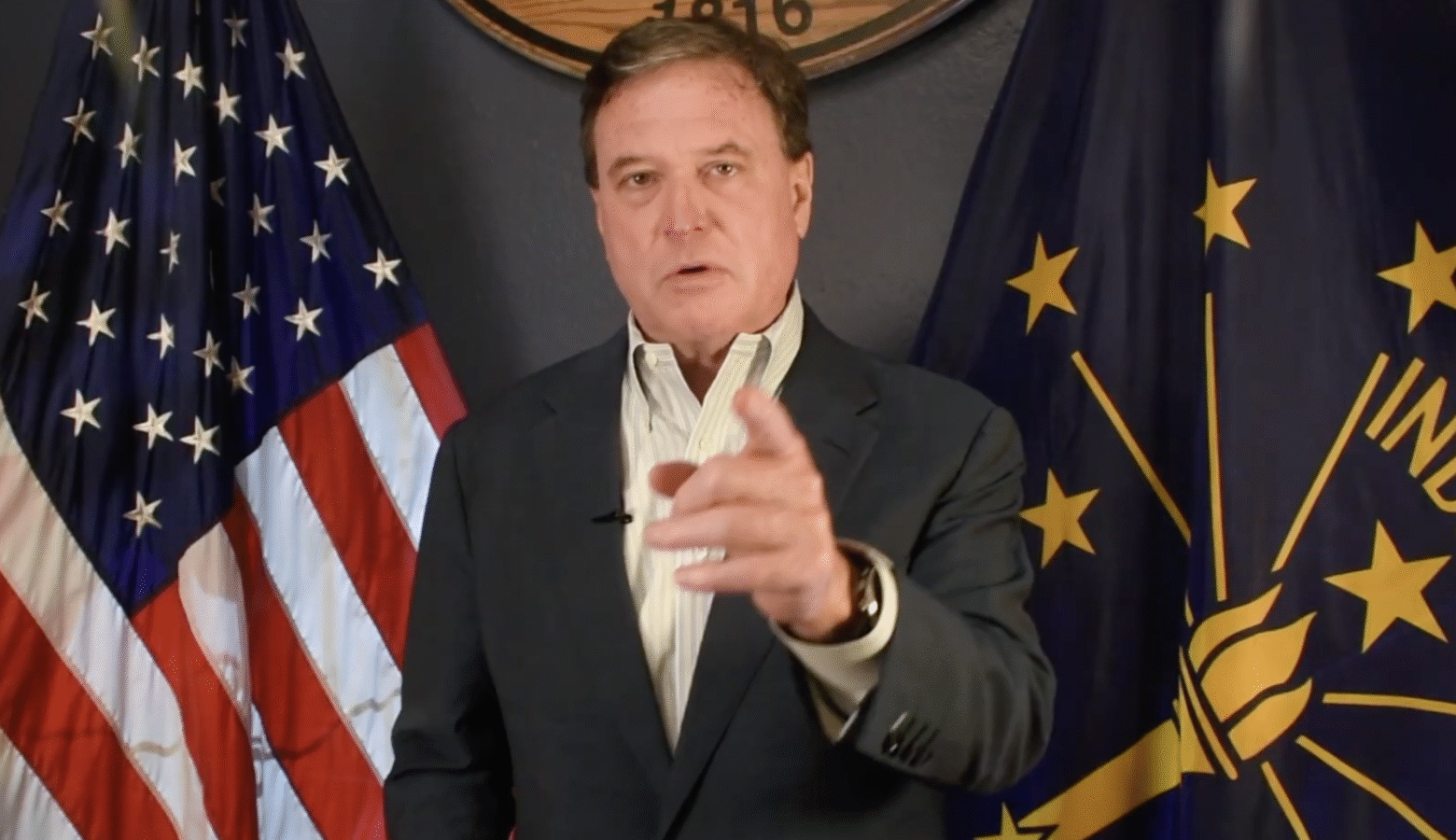A screenshot of a video shows Todd Rokita pointing at the camera, flanked by American and Indiana flags. Rokita is a White man with dark hair. He is wearing a black suit coat and white shirt.