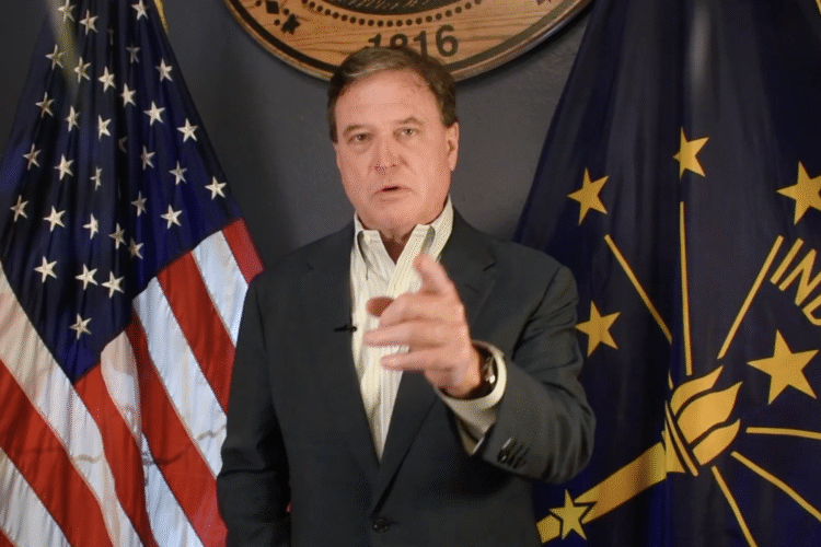 A screenshot of a video shows Todd Rokita pointing at the camera, flanked by American and Indiana flags. Rokita is a White man with dark hair. He is wearing a black suit coat and white shirt.
