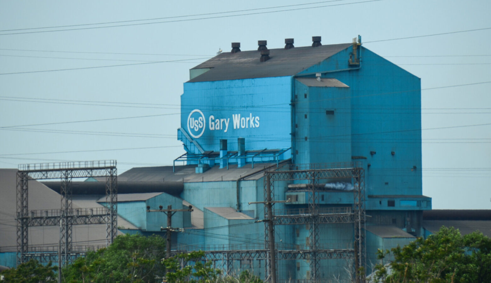 A tall, rusted blue building has the U.S. Steel logo with Gary Works written next to it in white letters. There are trees beneath the building.