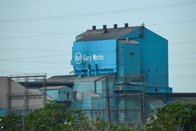 A tall, rusted blue building has the U.S. Steel logo with Gary Works written next to it in white letters. There are trees beneath the building.