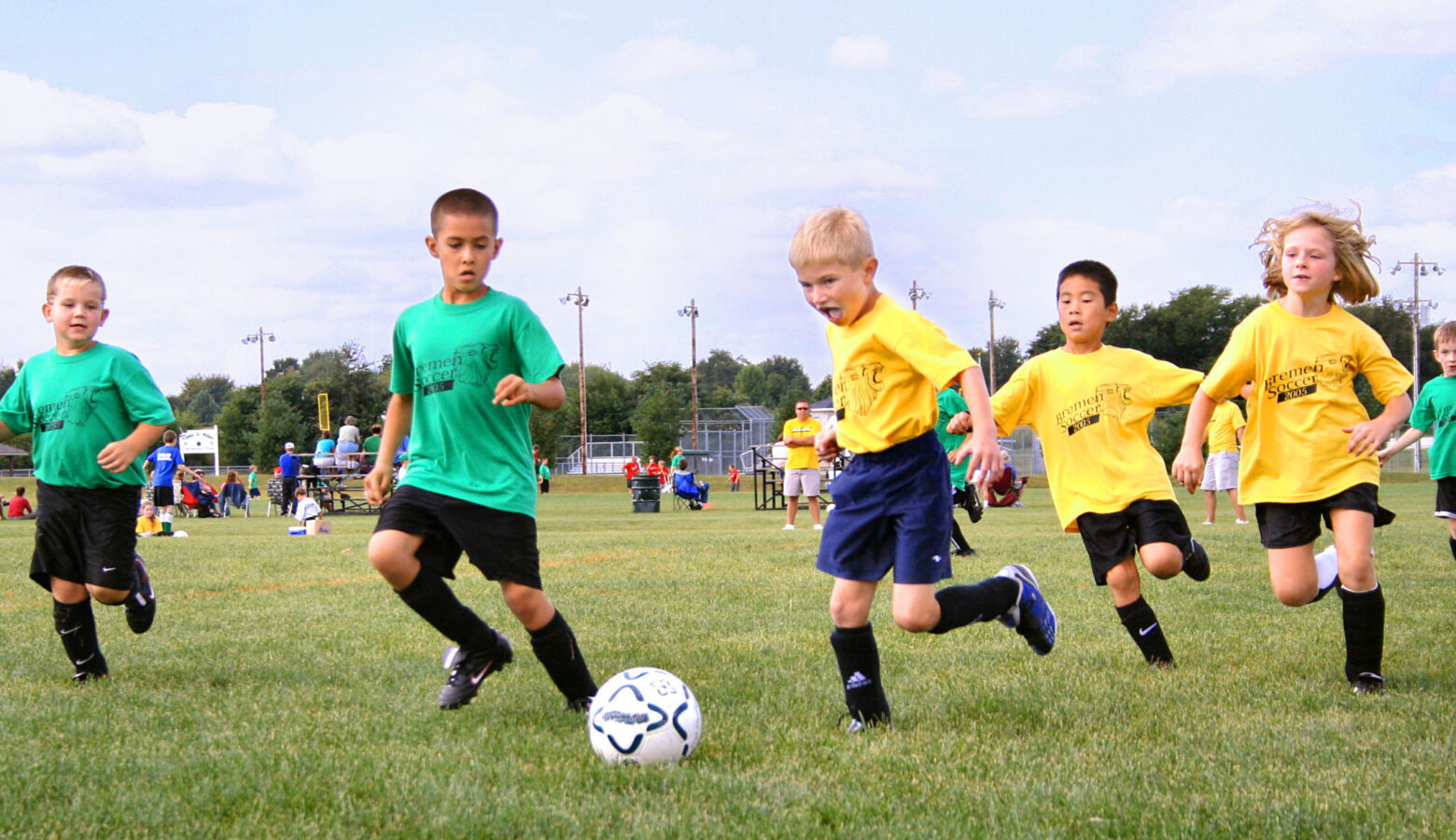 Children in green and yellow shirts run and kick a soccer ball.