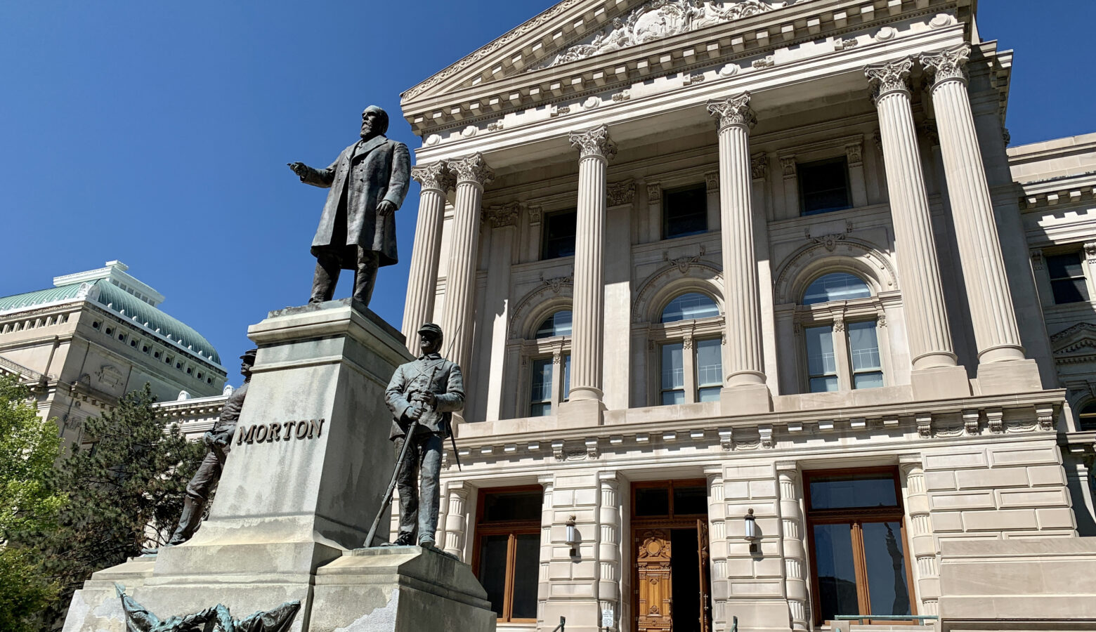 The eastern exterior of the Indiana Statehouse is four tall columns leading up to a triangular roof. The columns are above a large wooden doorway that's at the top of a set of stairs. In the foreground is a statute of former Governor Oliver Morton.