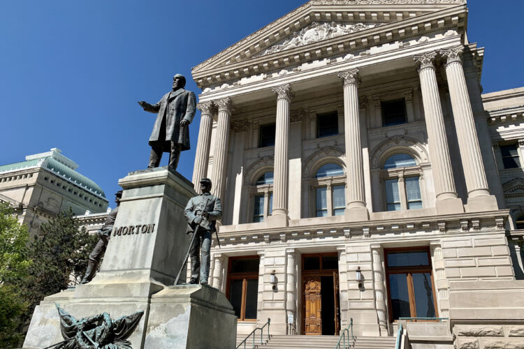 The eastern exterior of the Indiana Statehouse is four tall columns leading up to a triangular roof. The columns are above a large wooden doorway that's at the top of a set of stairs. In the foreground is a statute of former Governor Oliver Morton.
