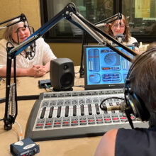 Two women sit in a studio chatting into microphones. We can see the shoulder of the woman they are talking to in the foreground, along with an audio soundboard between them.