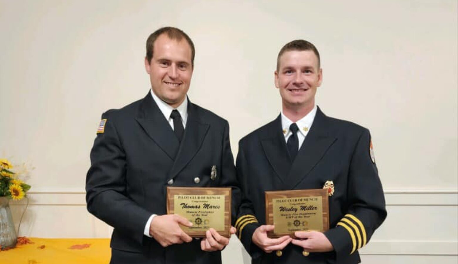 Two members of the Muncie Fire Department in dress uniform stand next to each other, holding gold plaques. Both are White men with short hair.