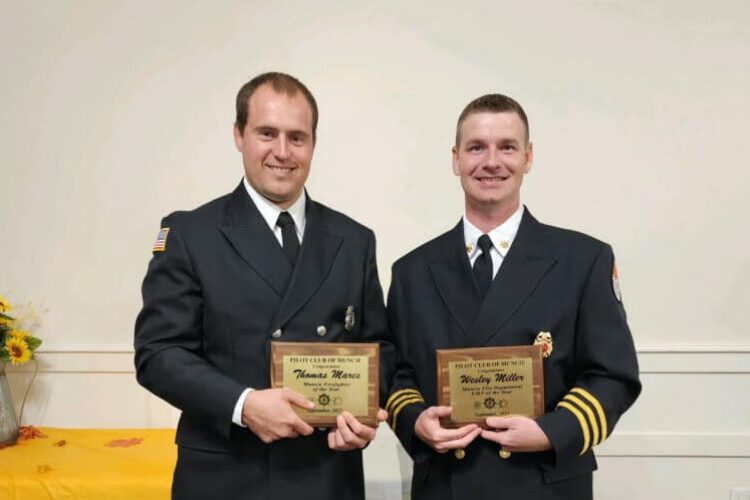 Two members of the Muncie Fire Department in dress uniform stand next to each other, holding gold plaques. Both are White men with short hair.