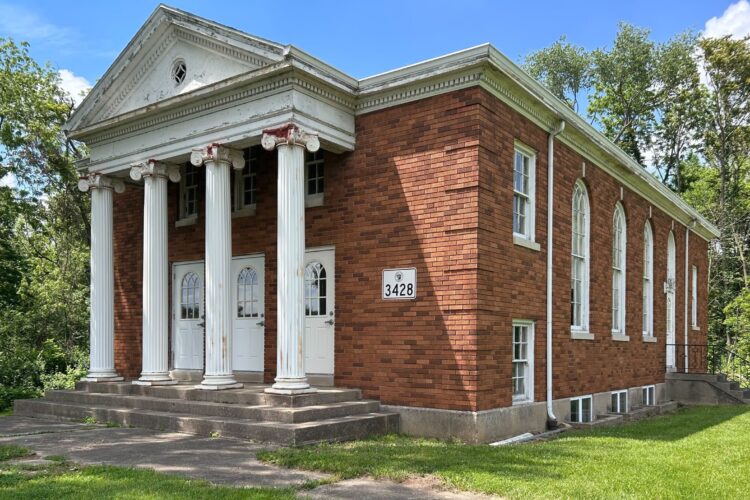 A brick building with old white pillars at its entrance sits in a grass field.
