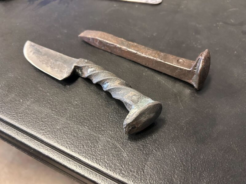 A rusty railroad spike sits next to a crude metal knife