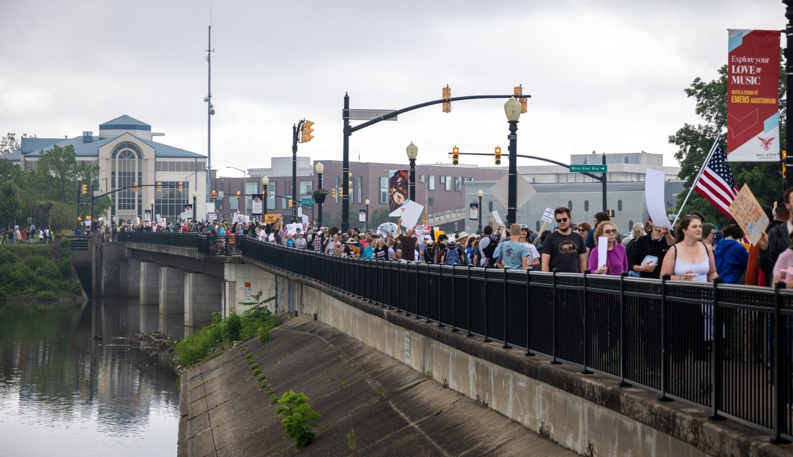 Hundreds of people waving signs and flags stand along a bridge, with the river below and the Muncie skyline in the background.