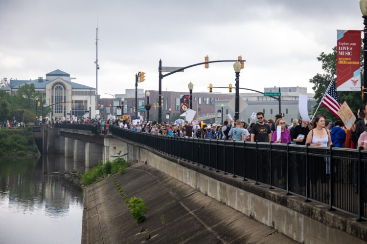 Hundreds of people waving signs and flags stand along a bridge, with the river below and the Muncie skyline in the background.