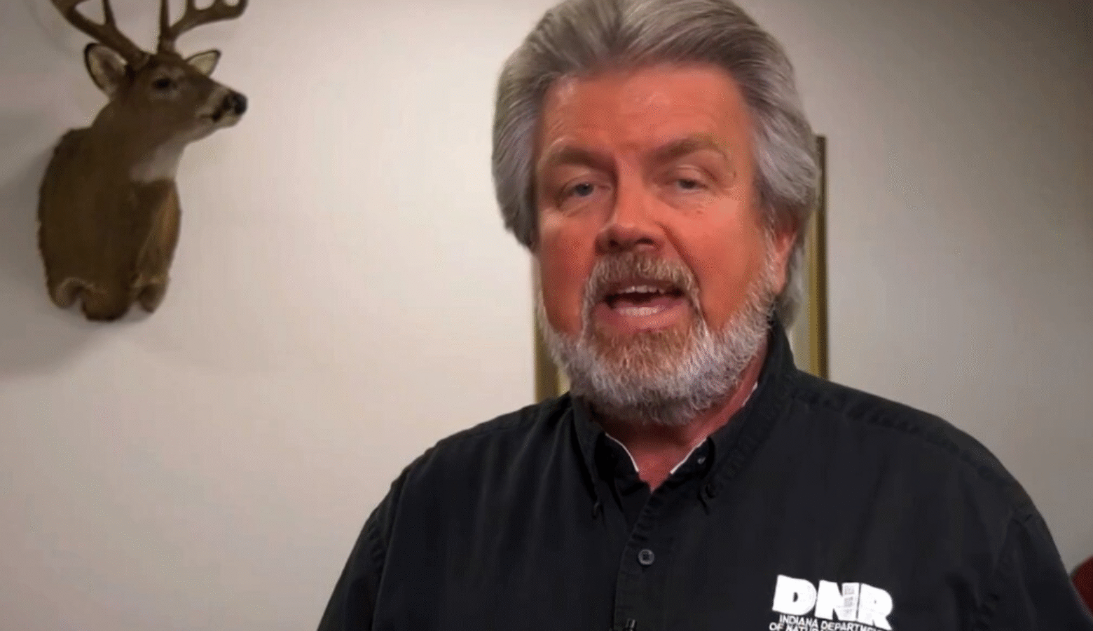 Former Indiana State Parks director and DNR director Dan Bortner. Bortner has medium length silver hair with a beard. He wears an Indiana DNR black polo. Behind him is a mounted on the wall is a deer head.