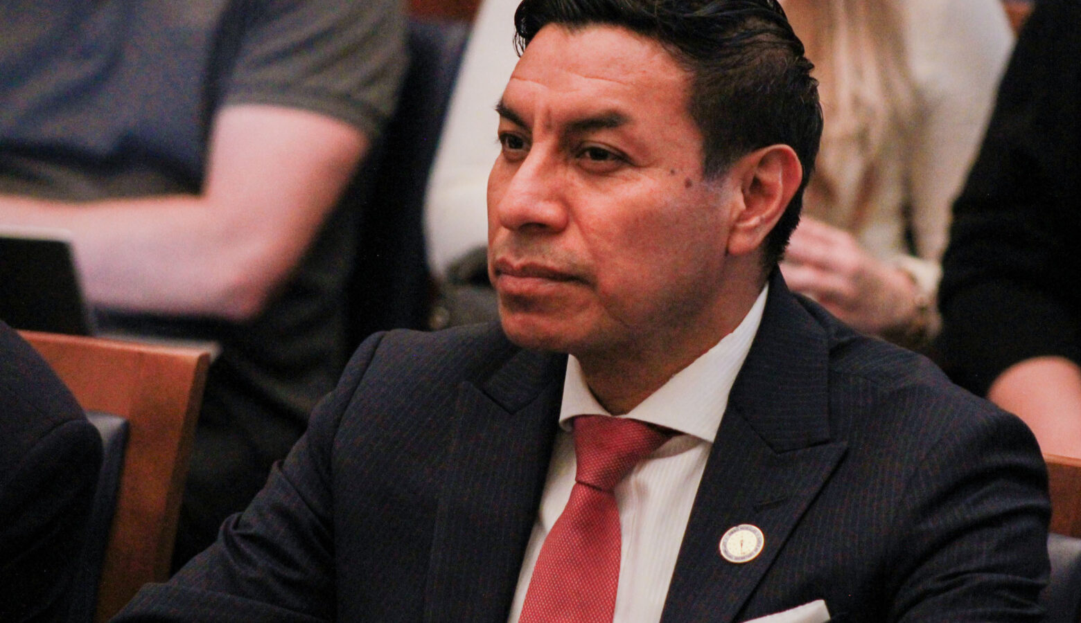 Diego Morales sits at a table and looks forward. Morales is a Latino man with black hair. He is wearing a dark suit, white shirt and red tie.