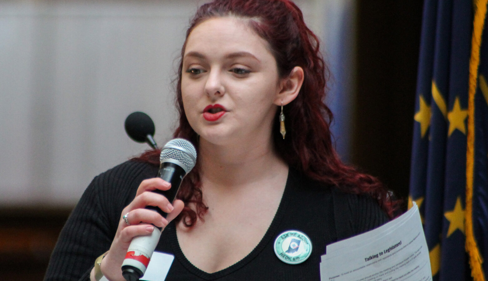 Eliza Brader speaks into a microphone at a rally in the Indiana Statehouse. She wears a black shirt and has her hair pulled back. Behind her is an Indiana state flag.