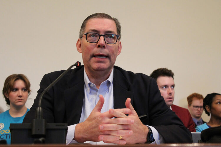 Family and Social Services Secretary Mitch Roob testifies in front of the Senate Appropriations Committee. He is seated at a wooden table, with a thin microphone on top of it that he's speaking into. He is a white man with short black hair that gets grayer as it goes down his head. He wears a plain black blazer, a slightly tinted white shirt with a single button undone, black glasses and a simple gold ring.