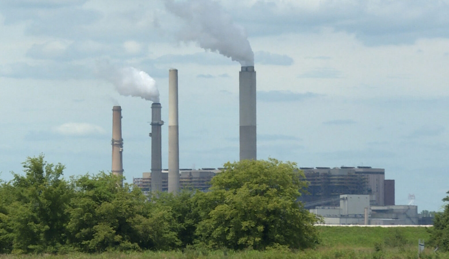 Four tall, long, gray and blue poles stand miles away from a tan building. The two blue poles are producing gray smoke from the top. Four green bushes are covering the bottom of the poles. The sky is cloudy with gray and blue clouds covering the area.