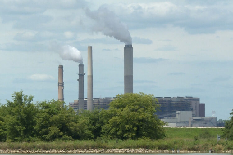 Four tall, long, gray and blue poles stand miles away from a tan building. The two blue poles are producing gray smoke from the top. Four green bushes are covering the bottom of the poles. The sky is cloudy with gray and blue clouds covering the area.