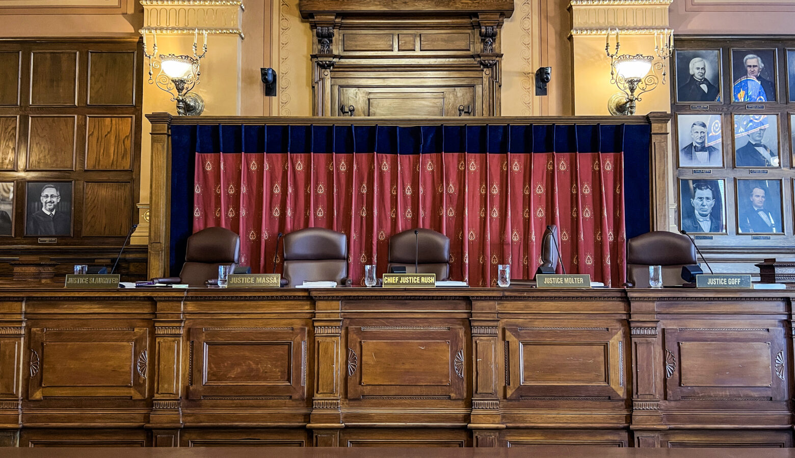 The Indiana Supreme Court chamber. There are five chairs at a long wooden desk. In front of each seat is a name plate with the name of each Indiana Supreme Court justice. Behind the chairs is a red curtain with gold symbols on it.
