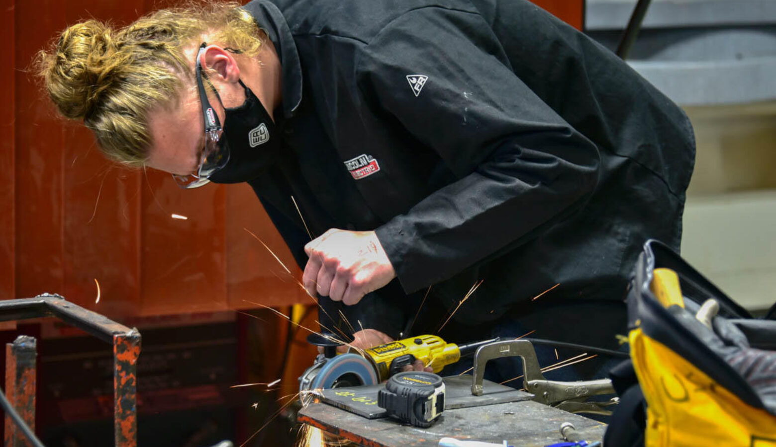 A young woman wears an all black outfit with a black mask. She has on clear safety goggles while she works on a project.