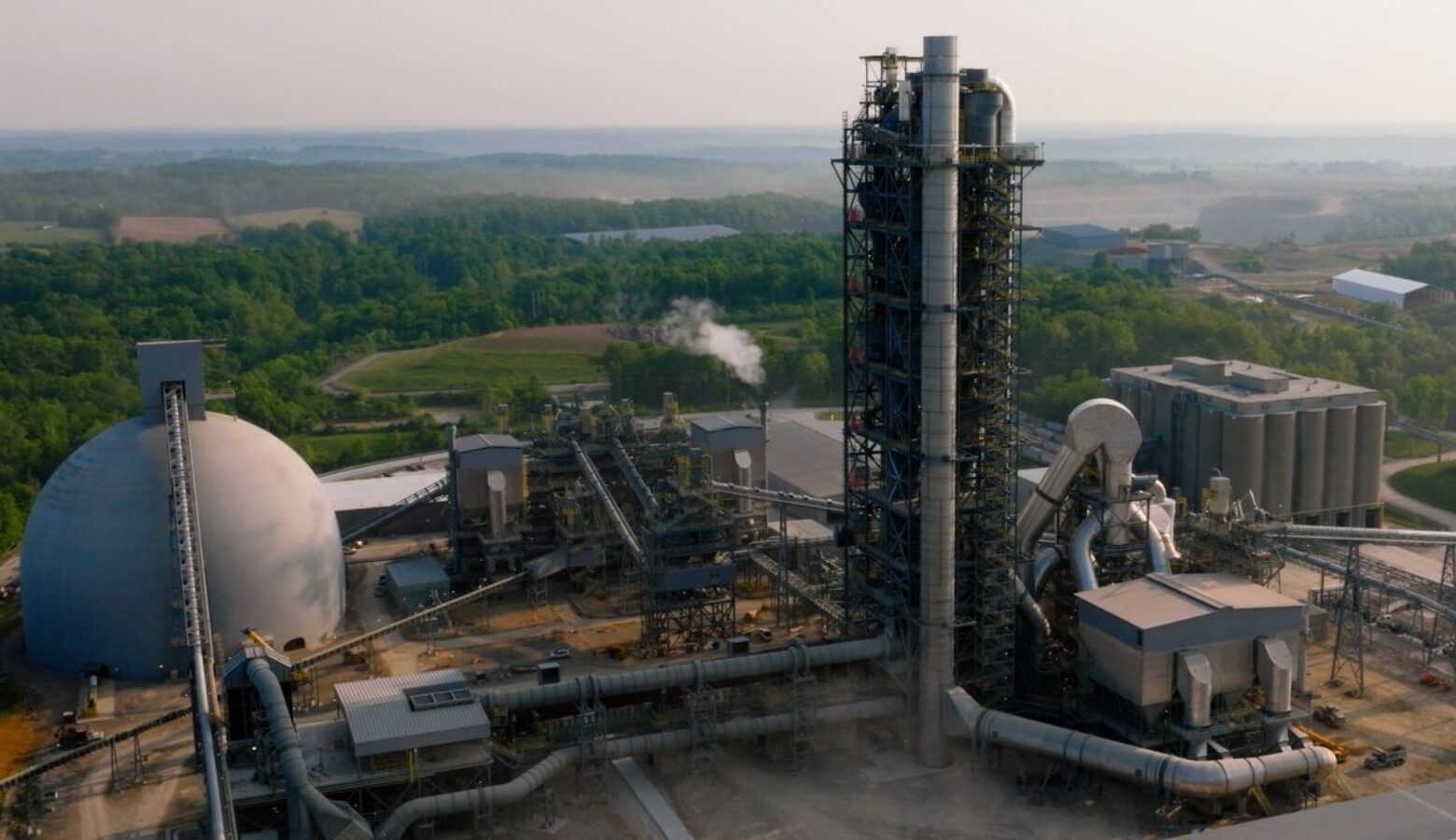An exterior wide shot of the Heidelberg cement plant in Mitchell. In the photo are a dome on the left of frame, numerous large pipes, a tall and wide pipe bedecked tower, and a block of silos on frame right.