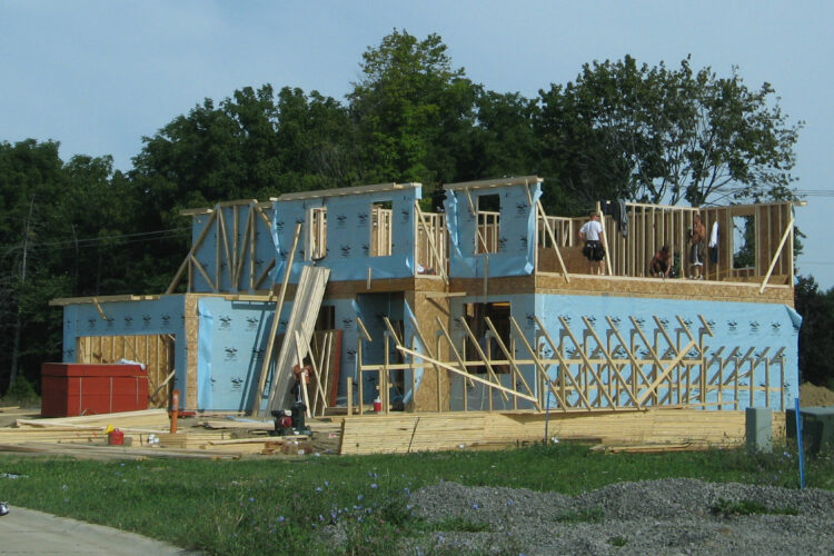 A new home under construction. It's a two story building. Some of the exterior walls are covered with blue sheets, with the rest of it exposed wood beams and walls.