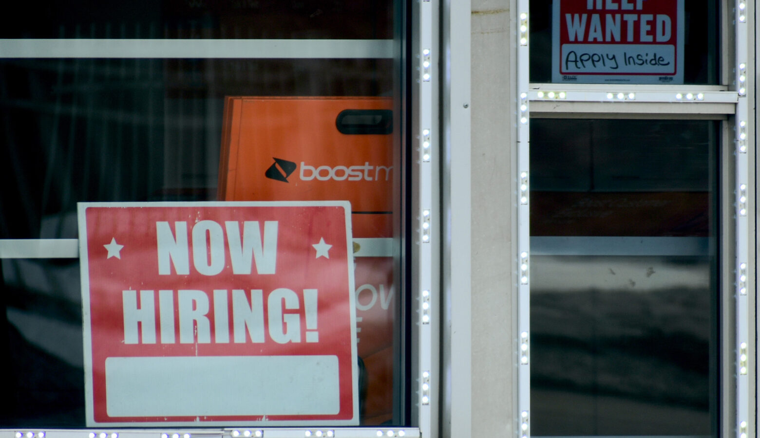 A red and white sign that said "Now Hiring" in white bold capital letters sits in the left lower half of a window with white borders. Behind the sign is a orange boost mobile box. to the upper right side of the sign is a smaller red and white sign that said "Help Wanted" in bold capital letters.