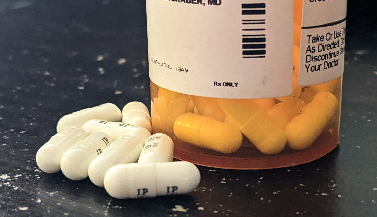 The bottom half of a prescription drug bottle has pills inside it visible below a white label with text on it. A few white capsules are outside the bottle.