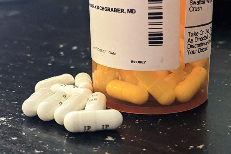 The bottom half of a prescription drug bottle has pills inside it visible below a white label with text on it. A few white capsules are outside the bottle.