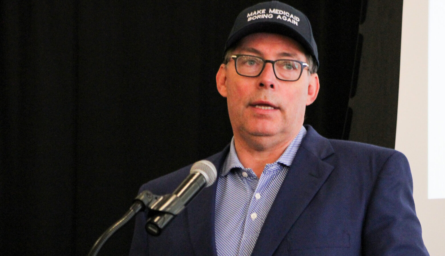 Mitch Roob, secretary of the Family and Social Services Administration, speaks into a microphone while wearing a "Make Medicaid Boring Again" hat.