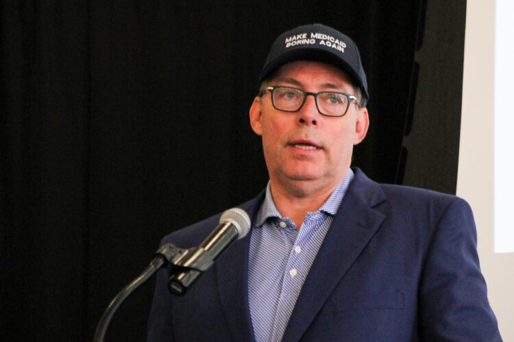 Mitch Roob, secretary of the Family and Social Services Administration, speaks into a microphone while wearing a "Make Medicaid Boring Again" hat.