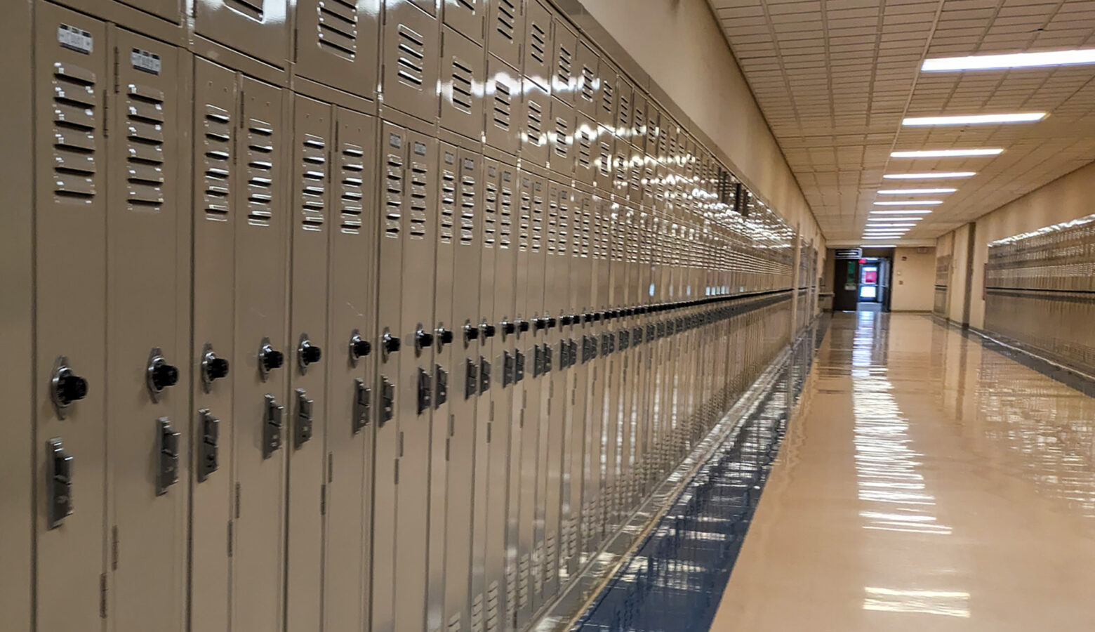An empty school hallway is lined by lockers. A digital clock at the top of the hallway reads the time and date. The fluorescent lights running down the hallway reflect off the linoleum flooring.
